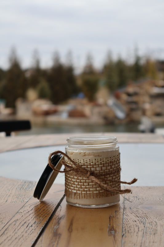 A glass jar of Sweet Lemon Whipped Shea Butter with a black metal lid leaning against its left side. In the background, a blurred rocky landscape with lush green trees adds a natural, earthy feel.